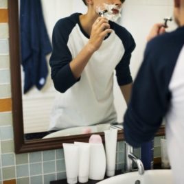 Boy wearing baseball shirt shaving in tiled bathroom