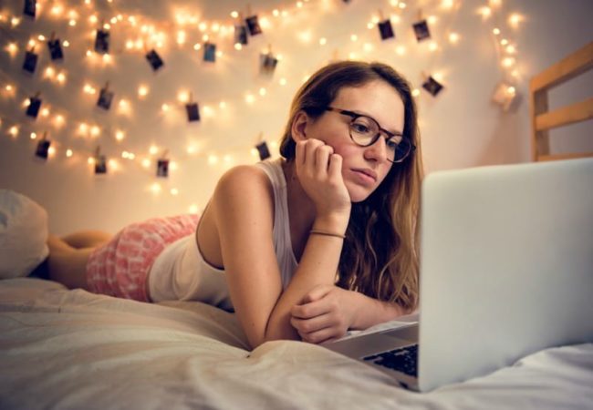 Dorm bed with white sheets and girl with glasses and laptop